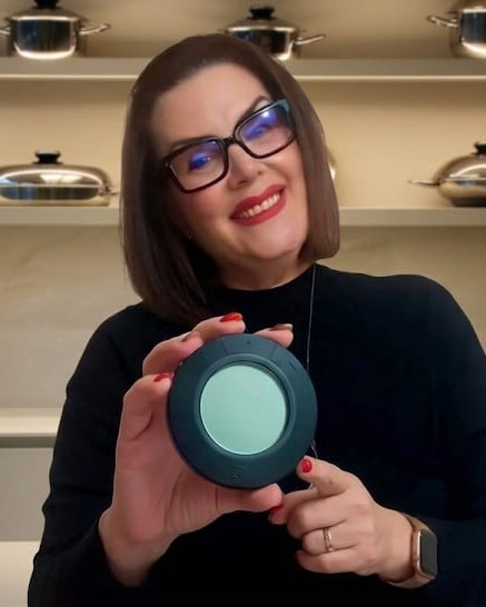 Charo Coromoto sonriendo en una cocina, con un temporizador en las manos y una estantería de ollas detrás