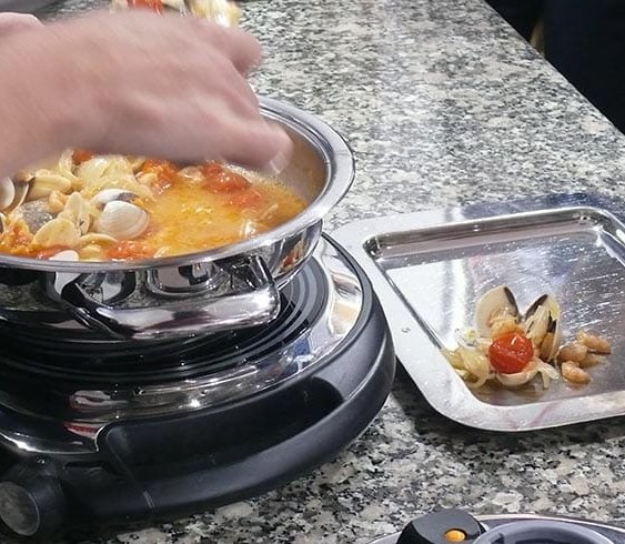 Una mano remueve almejas y tomates cherry en una sartén de acero sobre un fuego eléctrico, durante un taller de cocina