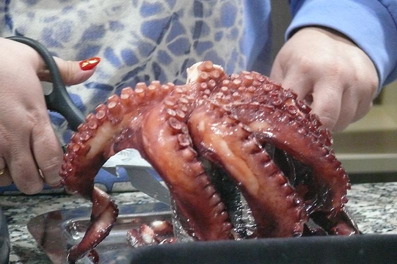 Unas manos cortan con tijeras un pulpo recién cocido, sostenido en vertical sobre una bandeja